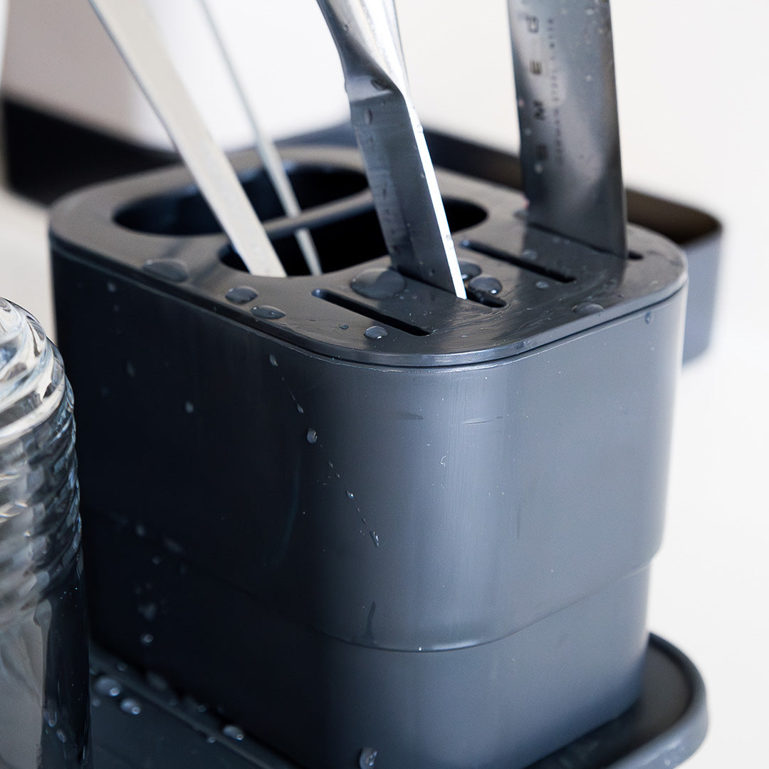 Escorredor de talheres moderno em plástico escuro com divisórias verticais, contendo utensílios metálicos molhados, com gotas de água visíveis na superfície.